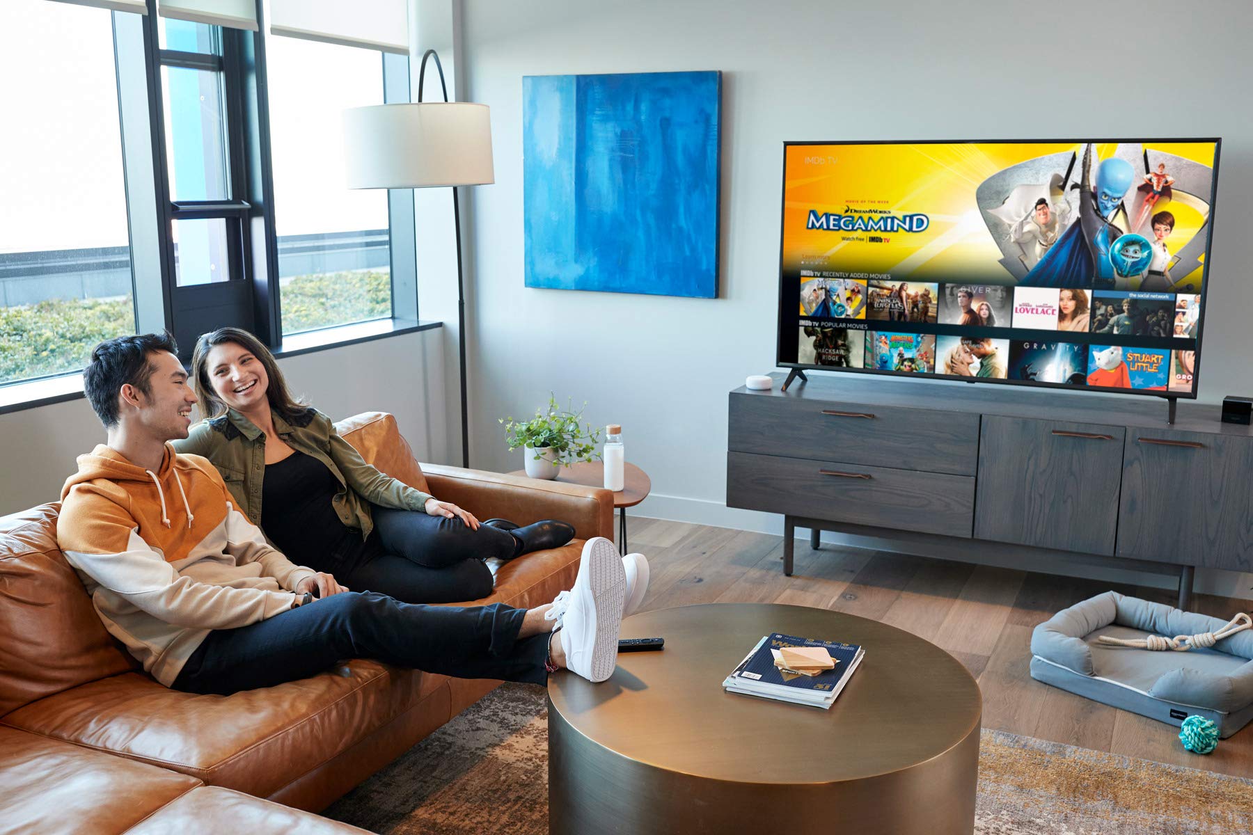 位年轻男子和一位年轻女子一起笑着坐在客厅沙发上,背景中有一台电视